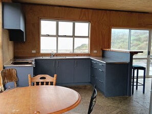 Een compacte keuken met een houten aanrecht, een spoelbak en kastjes. Een raam zorgt voor daglicht en uitzicht op de omgeving. Een ronde houten tafel met stoelen staat in het midden.