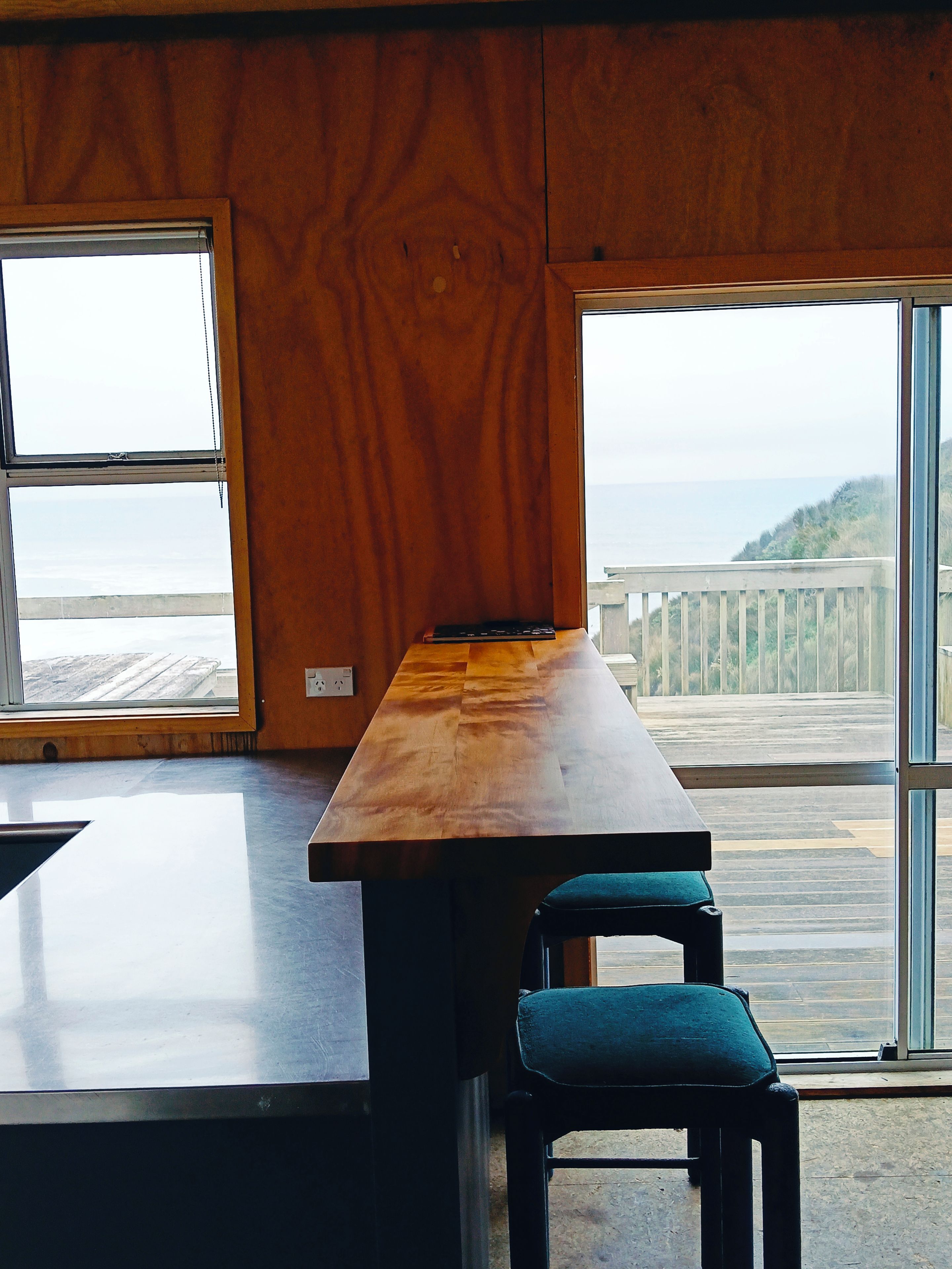 A room with a wooden table and a chair, large windows with a view of the sea and a wooden deck outside.