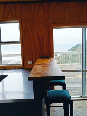 A room with a wooden table and a chair, large windows with a view of the sea and a wooden deck outside.