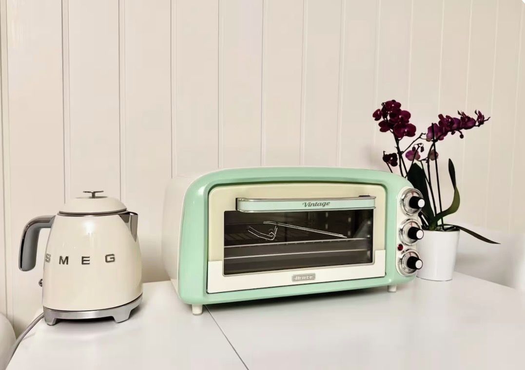 Um forno elétrico retrô e uma chaleira sobre uma superfície branca, com um vaso de planta ao lado.