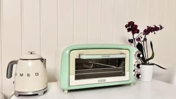 Um forno elétrico retrô e uma chaleira sobre uma superfície branca, com um vaso de planta ao lado.