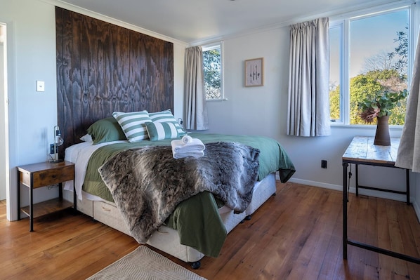 A bedroom with a bed, a nightstand, a bedside table, a window with curtains, and a view of trees. - The Jumping Trout Rustic Hideaway (Turangi)
