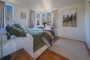 A bedroom with a bed, a bedside table, a lamp, a window with curtains, a painting on the wall, and a rug on the floor. - The Jumping Trout Rustic Hideaway (Turangi)