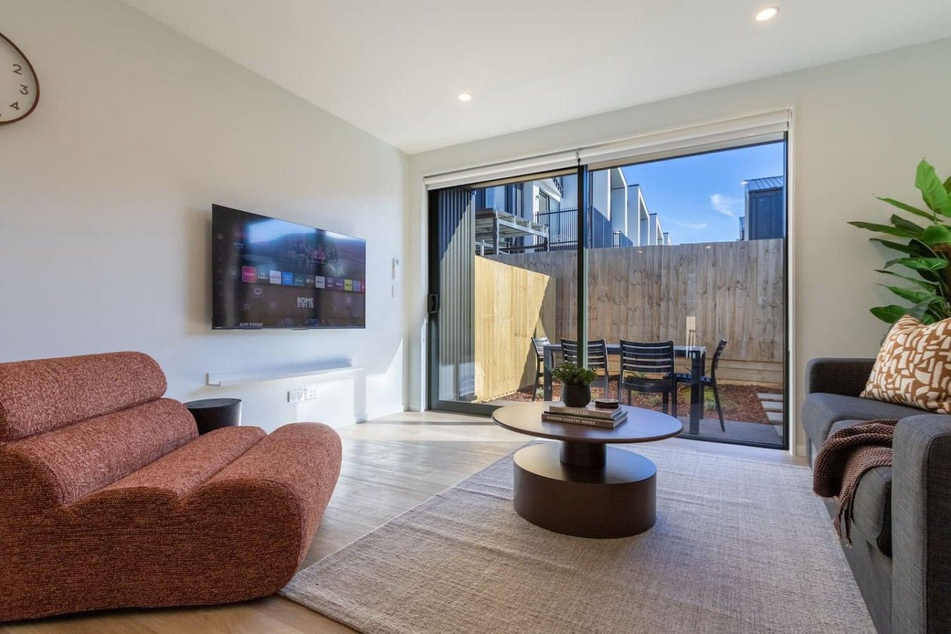 Uma sala de estar moderna com televisão de tela plana, sofá, mesa de centro e porta de vidro deslizante que dá para área externa com cerca de madeira e cadeiras.