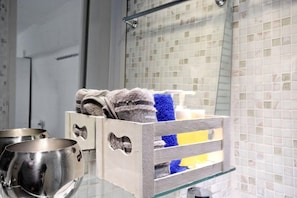 A bathroom with a glass shelf holding towels and toiletries, a metallic bowl, and a tiled wall. - Athens Plaza Luxury Apartments (Athens)