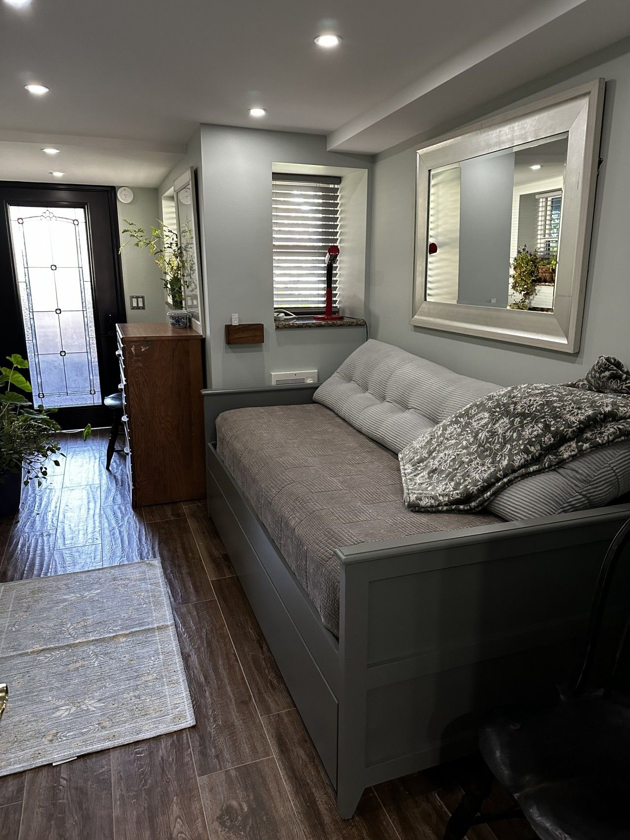 Une chambre à coucher comprenant un lit, une table de chevet, un miroir, une fenêtre avec des stores et une porte.