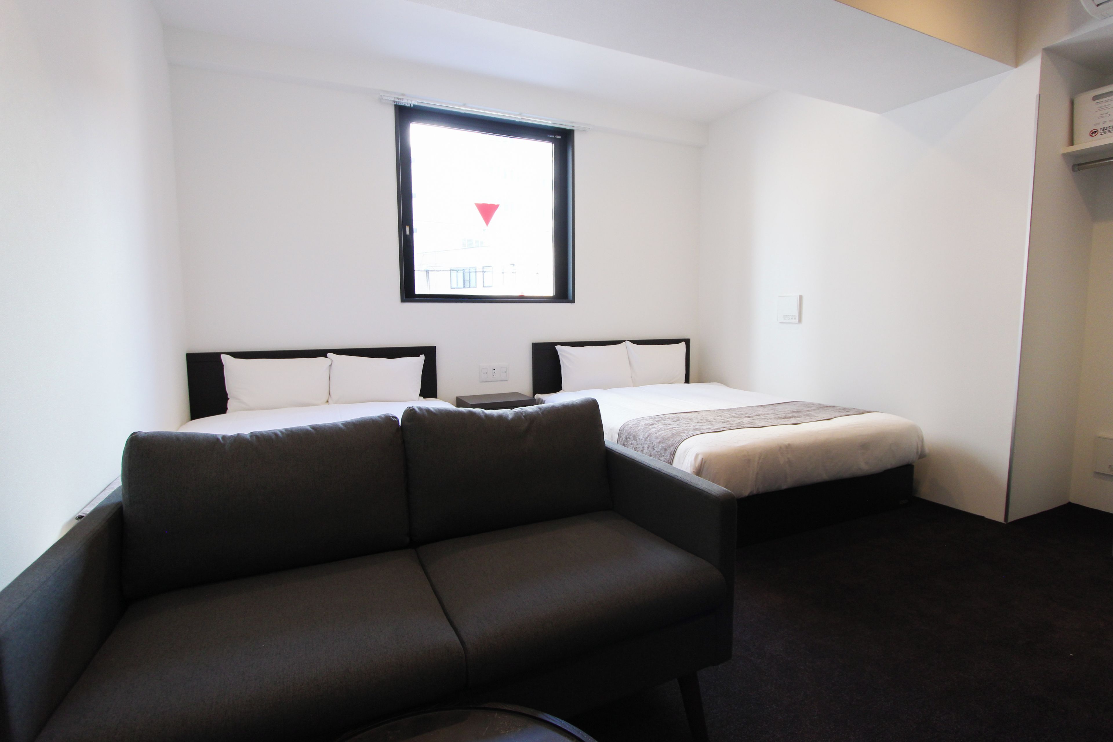 Habitació d'hotel petita amb un llit individual, un sofà, una taula i una finestra amb vista a la ciutat.