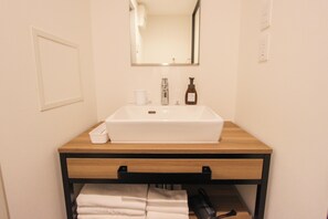 Un baño con un lavamanos blanco, un espejo, un perchero para toallas y una taza blanca pequeña.