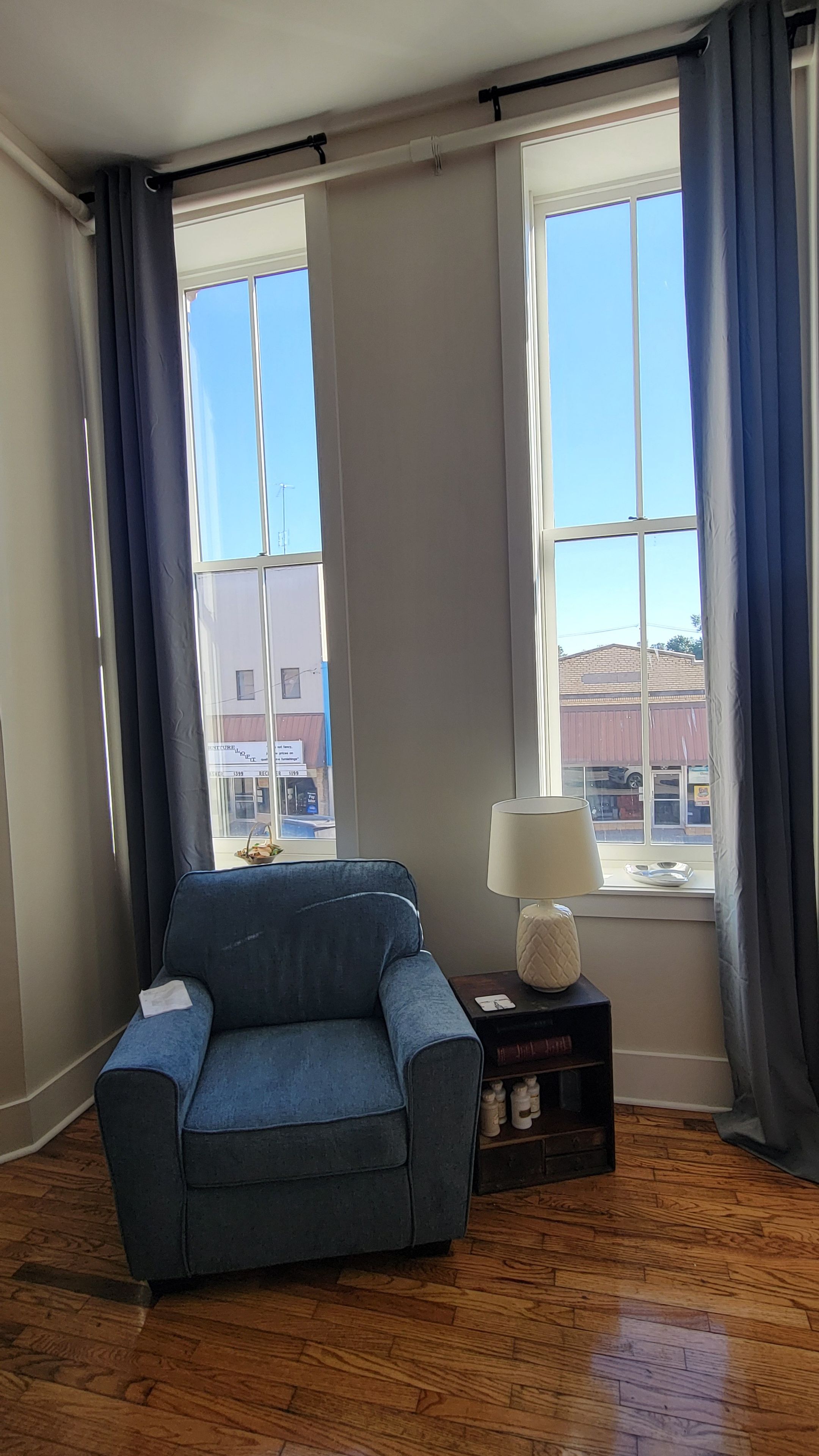 Une pièce avec un fauteuil bleu, une lampe sur une table d’appoint et de grandes fenêtres avec des rideaux.