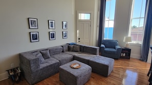 A living room with a sectional sofa, ottoman, framed pictures on the wall, a door, and a window with curtains.