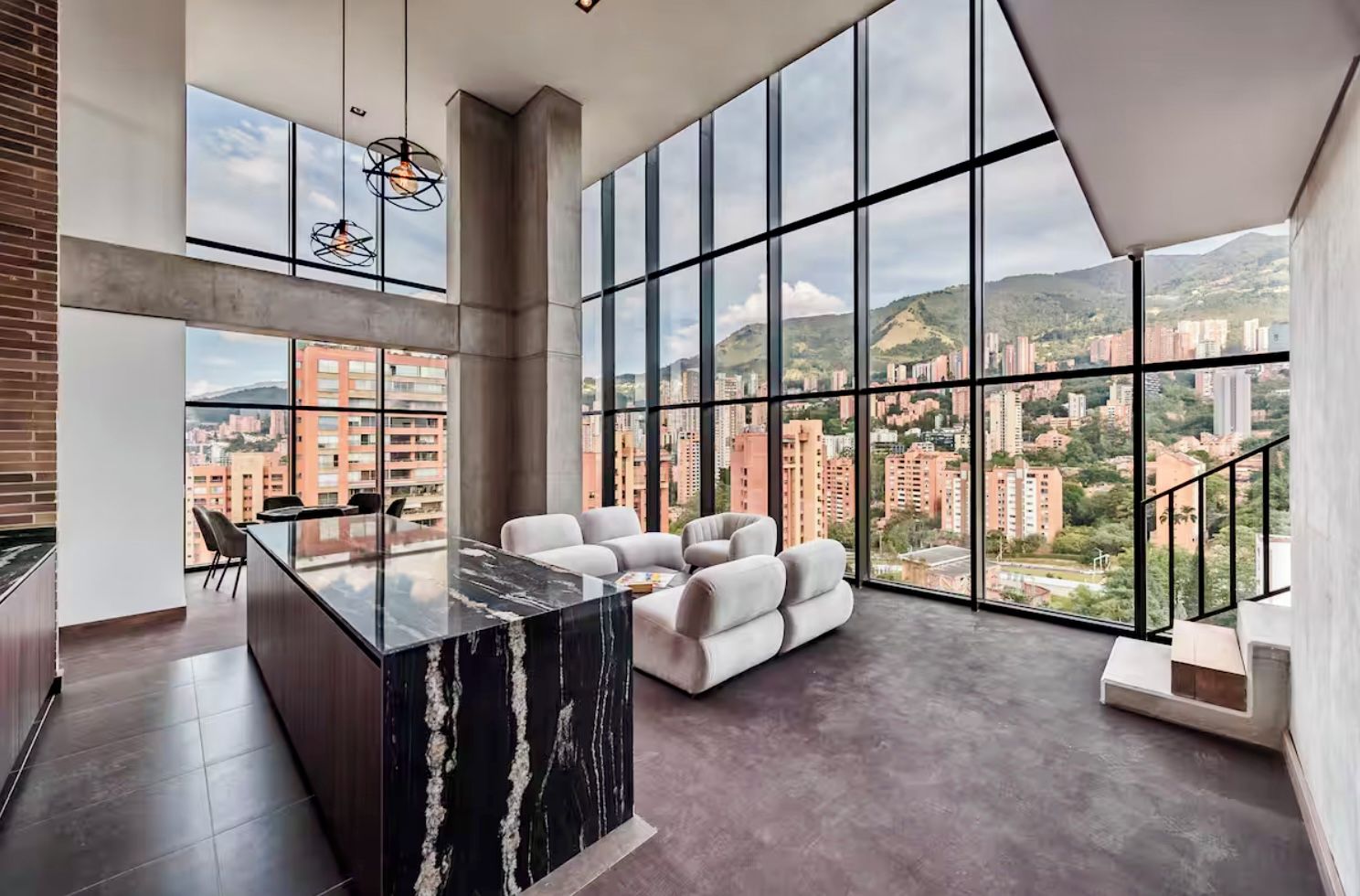 A modern interior with a marble kitchen island, a seating area with a sofa, and large windows offering a city view.