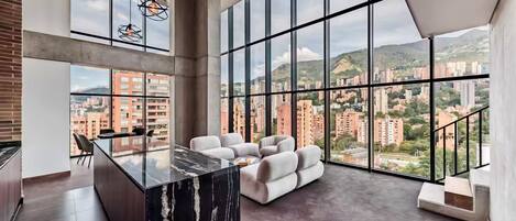 A modern interior with a marble kitchen island, a seating area with a sofa, and large windows offering a city view.