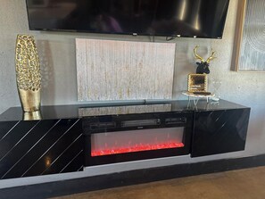 A black entertainment unit with a TV mounted above it, a decorative glass panel, and a fireplace with red flames. - Cozy 2-bedroom house in Idabel, OK only 10 miles to Broken Bow, OK (Idabel)