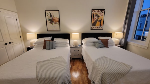 Habitación de hotel con dos camas, mesitas de noche con lámparas, un cartel enmarcado en la pared y una ventana con cortinas.