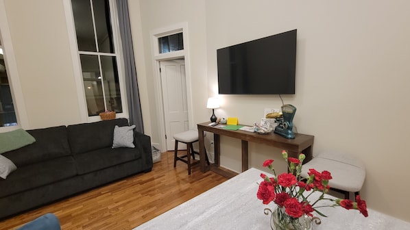 Un salon avec un canapé de couleur sombre, une petite table avec une lampe, un téléviseur fixé au mur et un vase rempli de fleurs rouges.