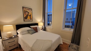 Habitación de hotel con cama, dos lámparas de noche, una mesita de noche, un reloj, un cartel enmarco y una ventana con vista a edificios.