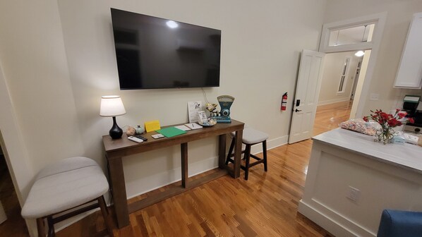 Una habitación con una televisión de pantalla plana montada en la pared, un escritorio de madera con una lámpara, una mesita y una silla.