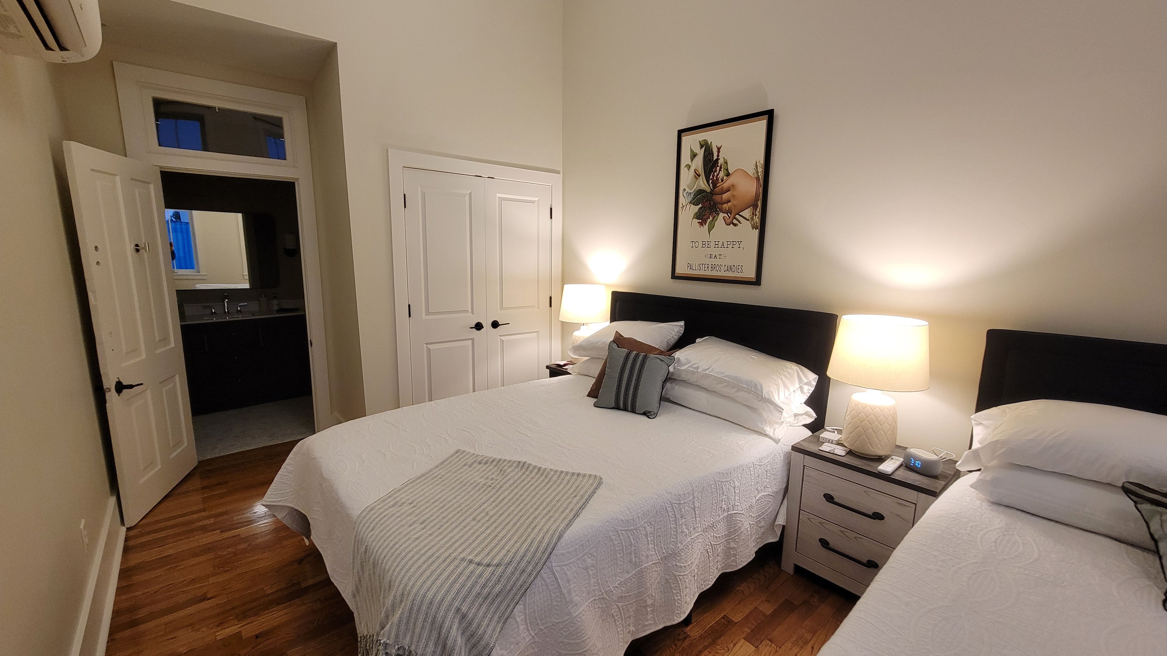 Ein Hotelzimmer mit einem Bett, Nachttischen, einer Wandleuchte, einem gerahmten Bild und einer Tür zu einem Badezimmer.