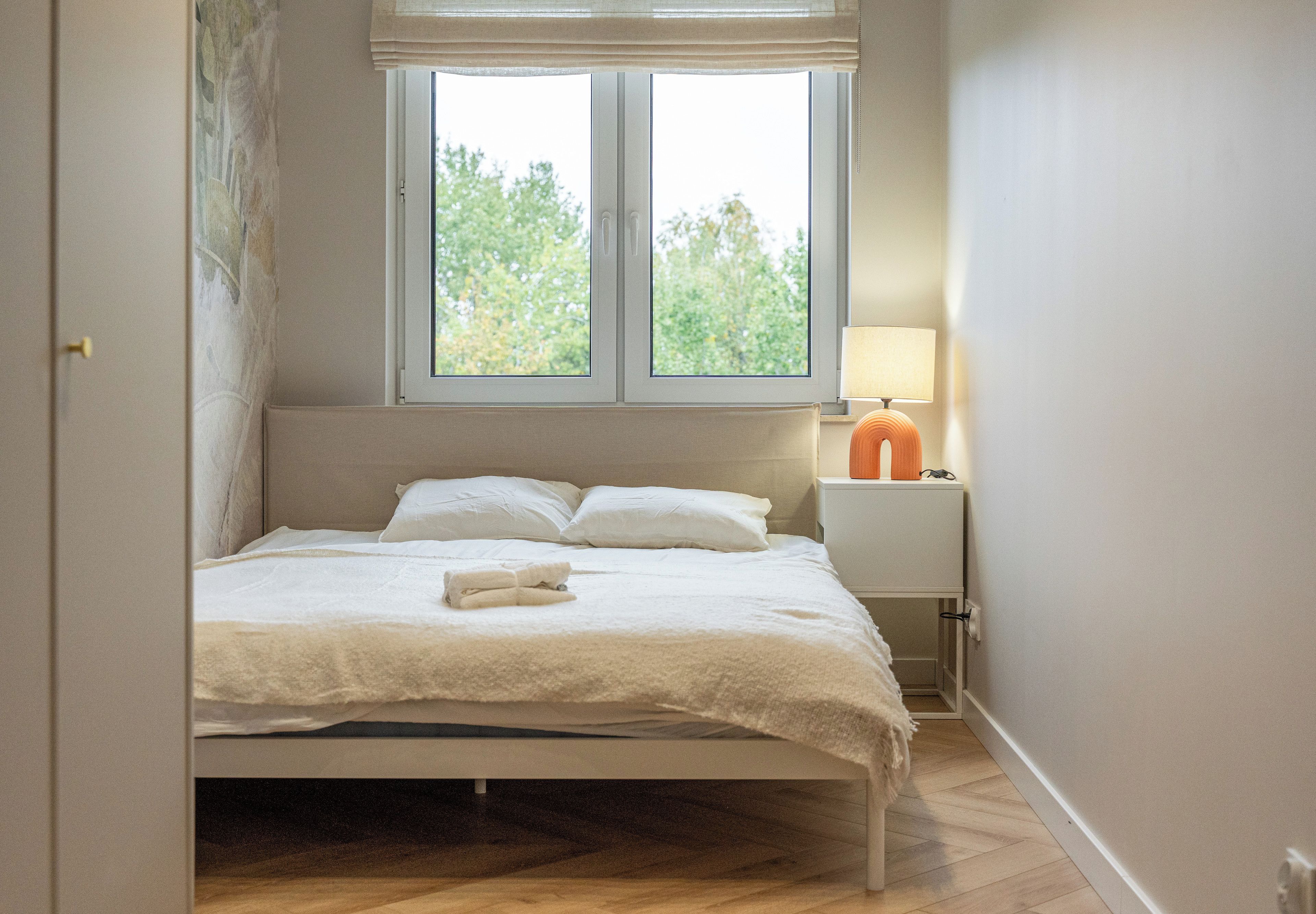 A bedroom with a bed, a nightstand, a lamp, and a window with a view of trees.