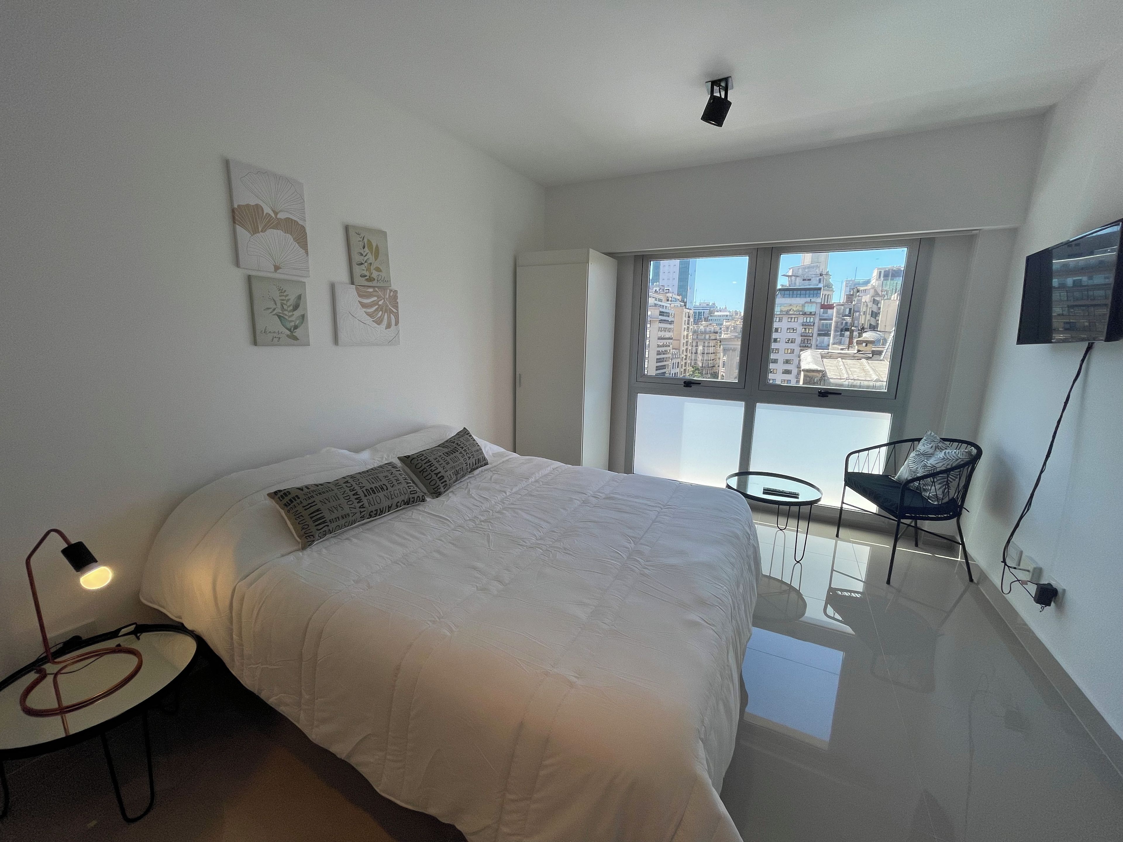 Un dormitorio moderno con una cama grande, una mesita redonda pequeña y una silla. Hay una ventana con vista a la ciudad y un televisor de pantalla plana montado en la pared.