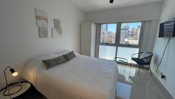 Un dormitorio moderno con una cama grande, una mesita redonda pequeña y una silla. Hay una ventana con vista a la ciudad y un televisor de pantalla plana montado en la pared.