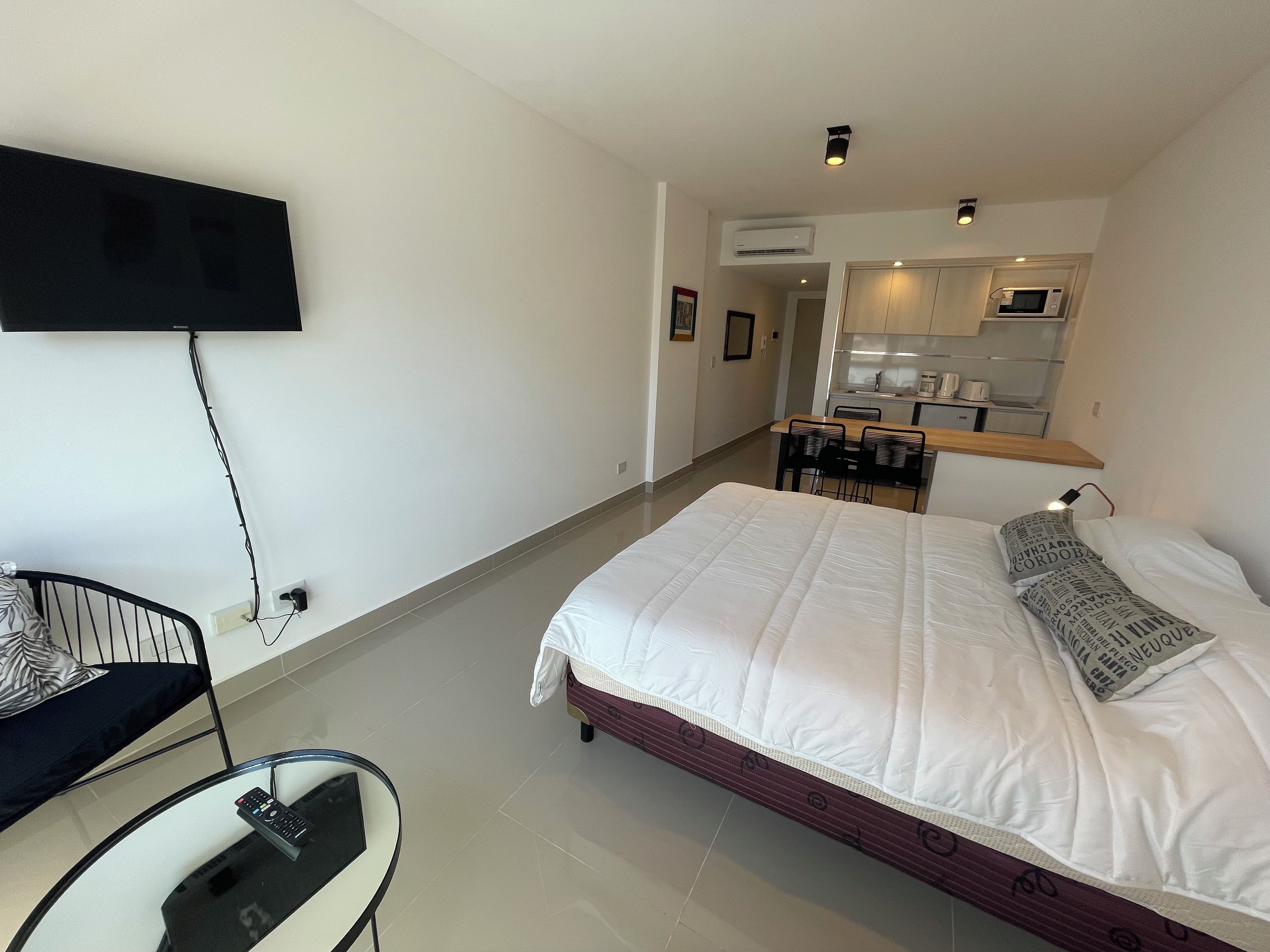 Habitación de hotel con cama, televisor montado en la pared, escritorio con silla y una kitchenette al fondo.