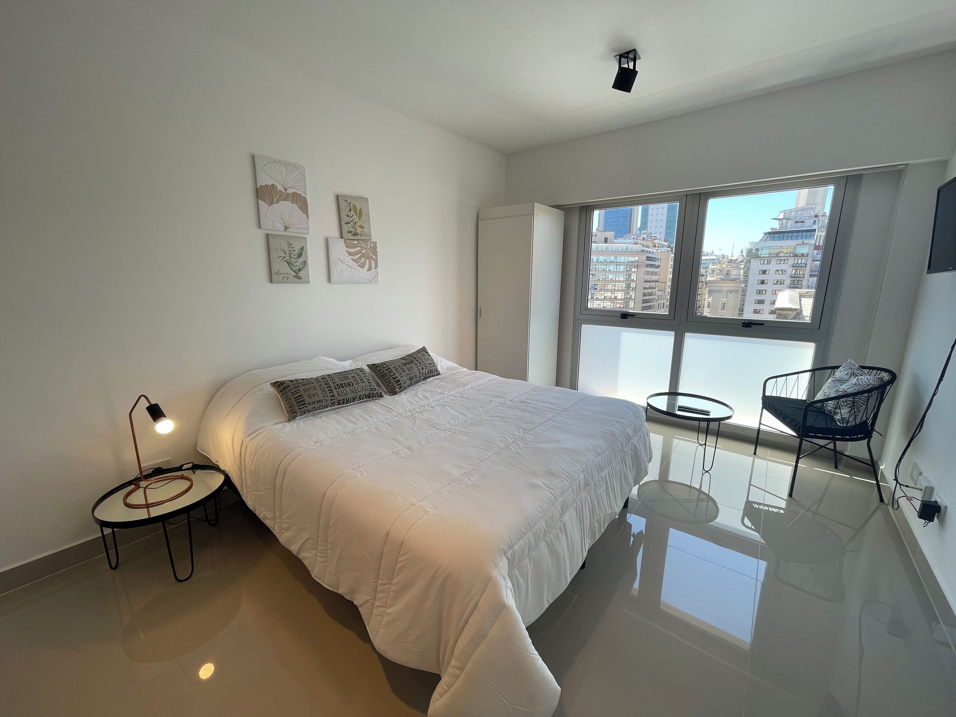 Un dormitorio moderno con una cama grande, una mesita de noche, una silla y una ventana con vista a la ciudad.