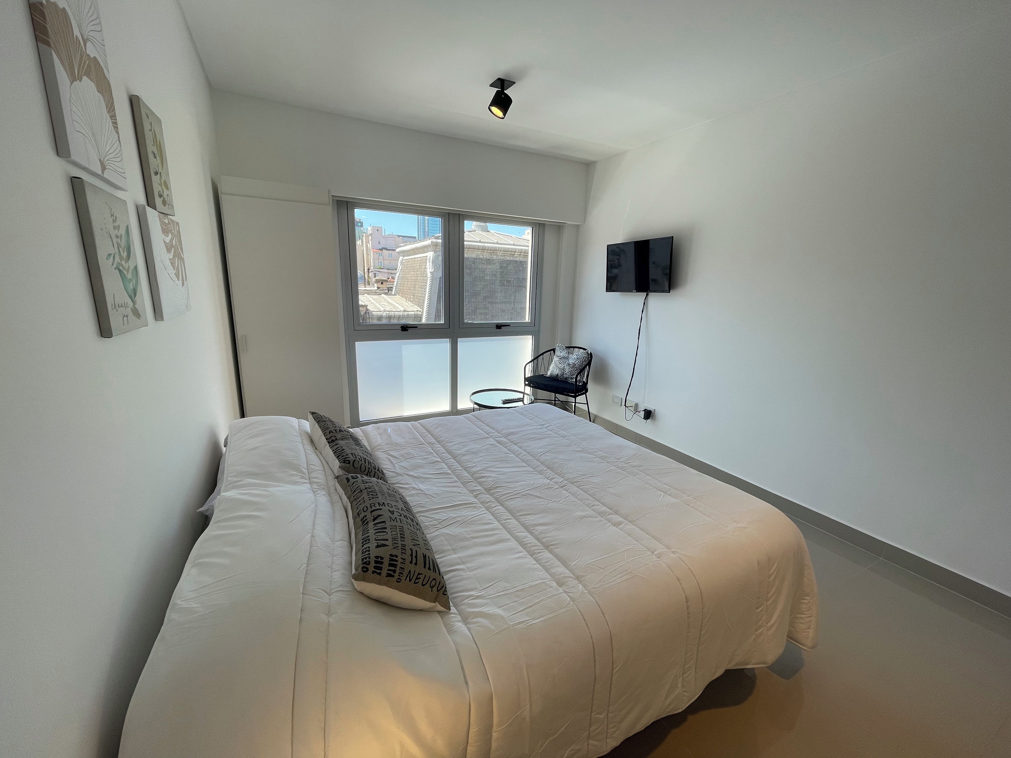 Un dormitorio con cama, una silla, televisor y ventana con vista a la ciudad.