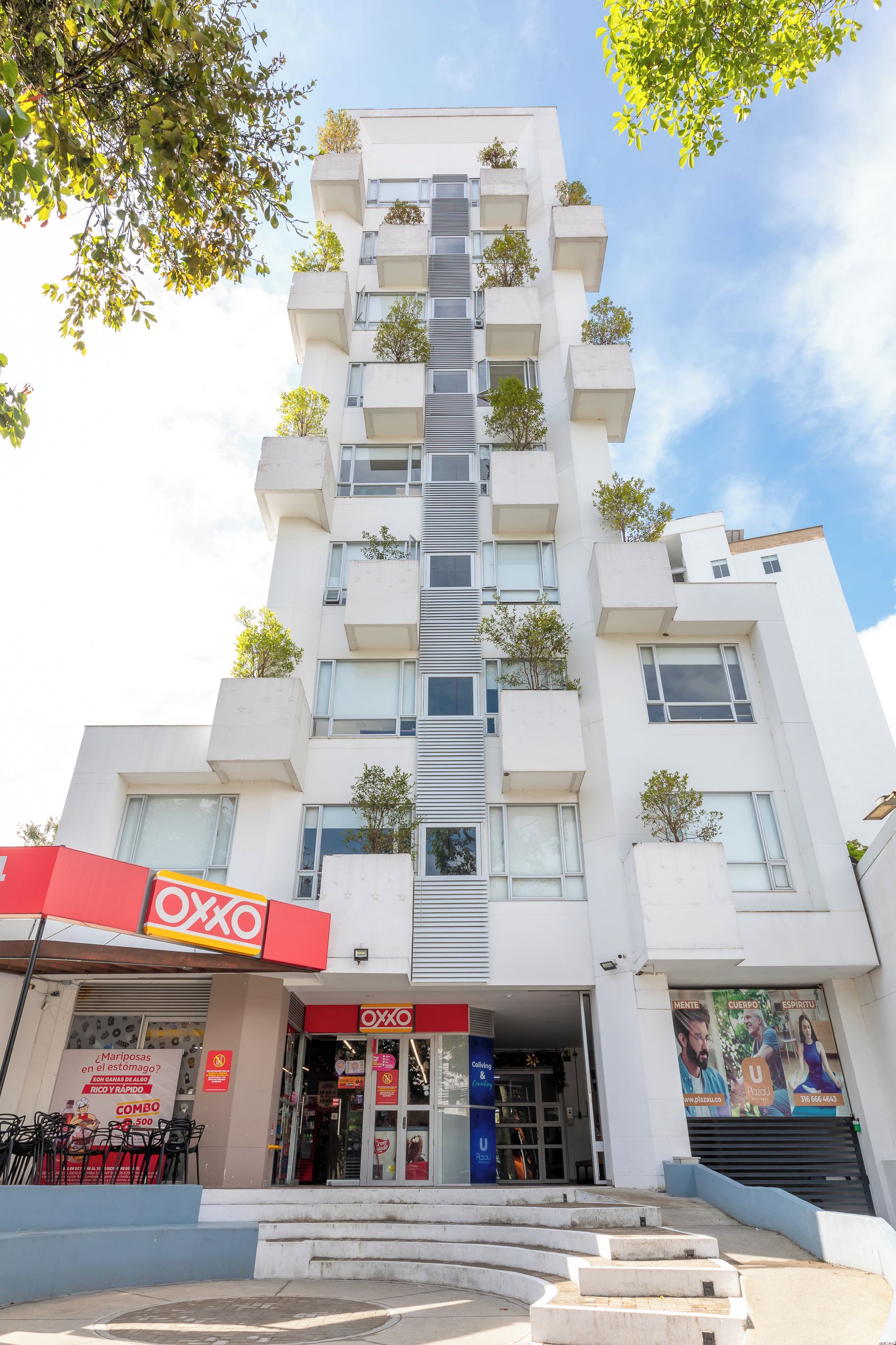 Un edificio de varios pisos con un diseño arquitectónico moderno, que cuenta con balcones con vegetación, un escaparate con señalización y una escalera que conduce a la entrada.