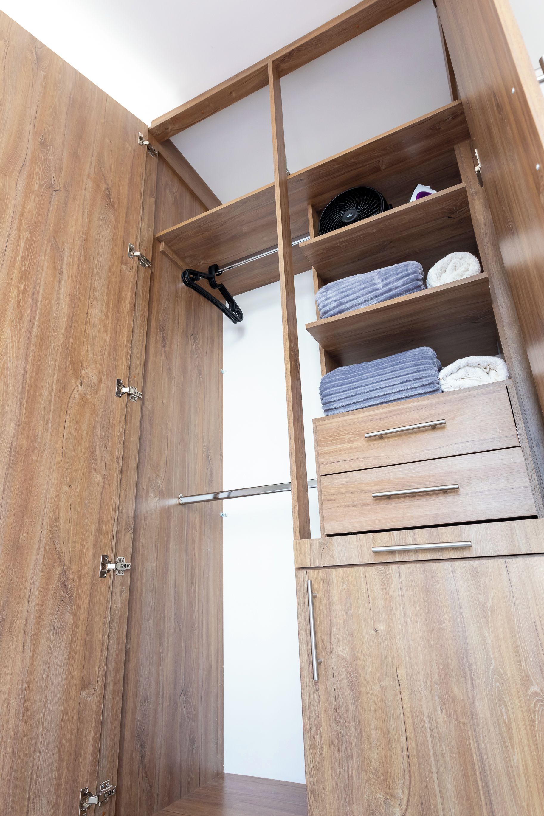 Un armario de madera con varios cajones, estantes y una barra para colgar, además de un ventilador.