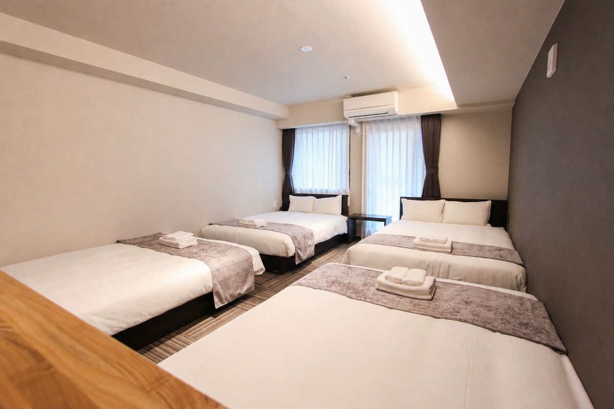 A hotel room with four single beds, a small bedside table, an air conditioning unit, and a window with curtains.