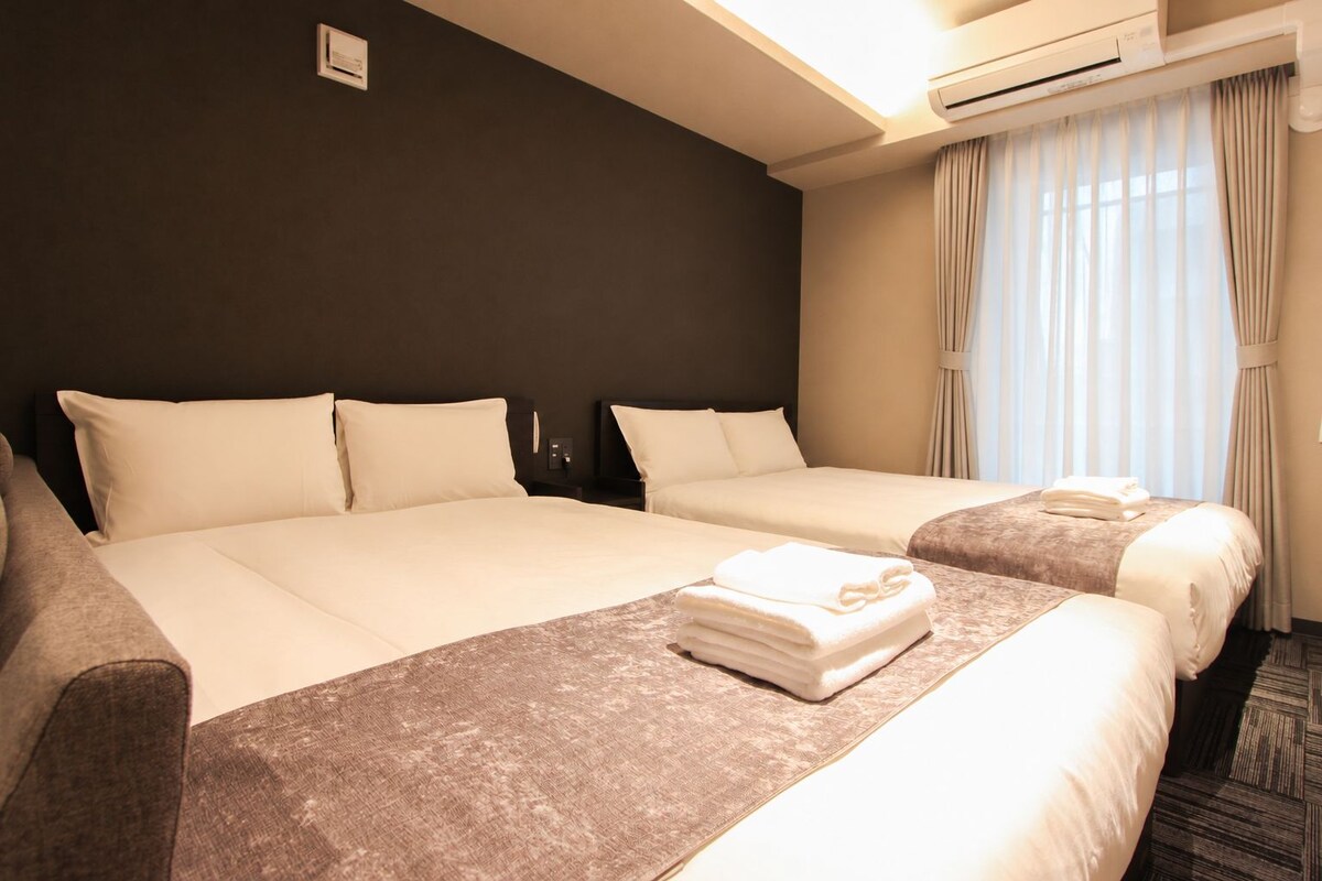 A hotel room with a large bed, a smaller bed, and a desk with a chair. There is an air conditioning unit above the bed and a window with curtains.