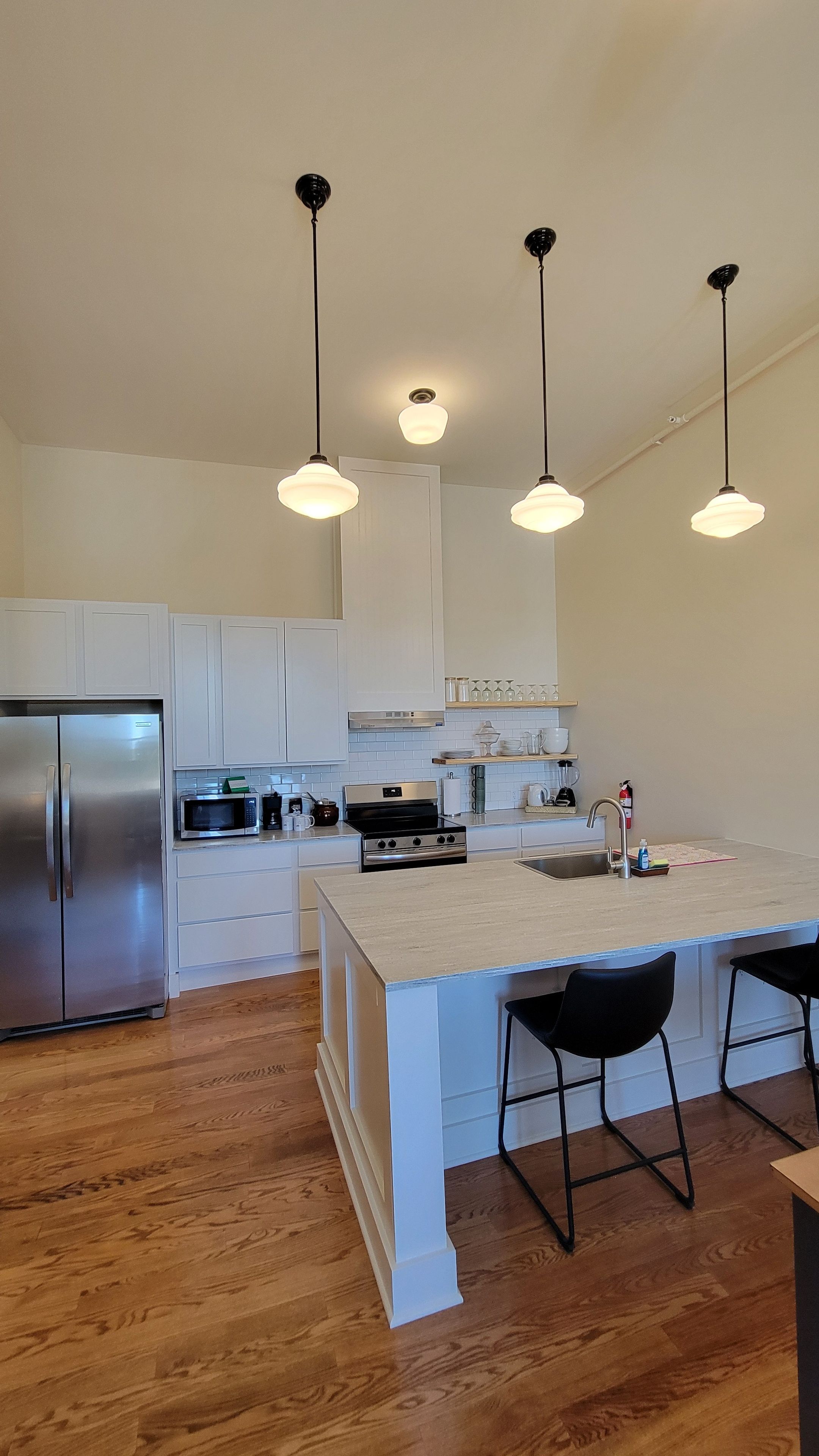 Una cocina moderna con armarios blancos, electrodomésticos de acero inoxidable, una isla central con taburetes y luminarias colgantes.