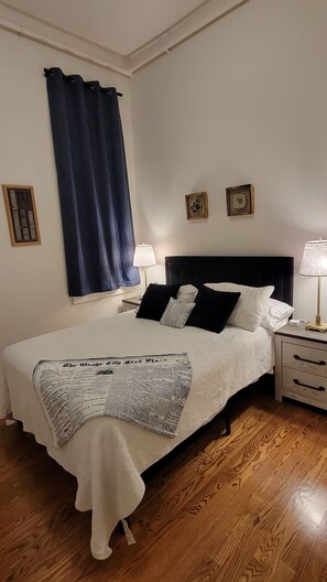 Una cama bien hecha con edredón blanco y una manta de periódico, una lámpara de noche y una mesita de noche con lámpara.