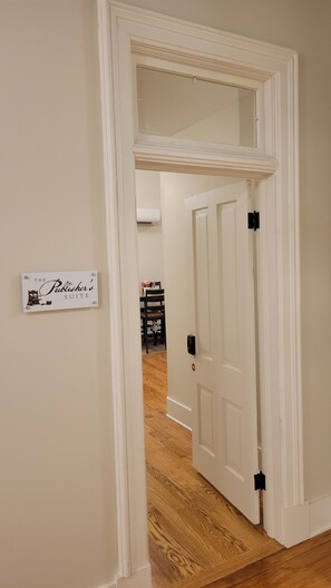 Ein Flur mit einem Schild, auf dem 'The Publisher's Suite' steht, der zu einem Zimmer mit Holzboden und einer Tür mit schwarzem Griff führt.