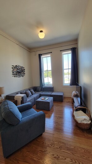 Ein Wohnzimmer mit Ecksofa, Couchtisch und Stehlampe. Zwei Fenster mit Vorhängen, eine Wandleuchte und ein Holzboden.