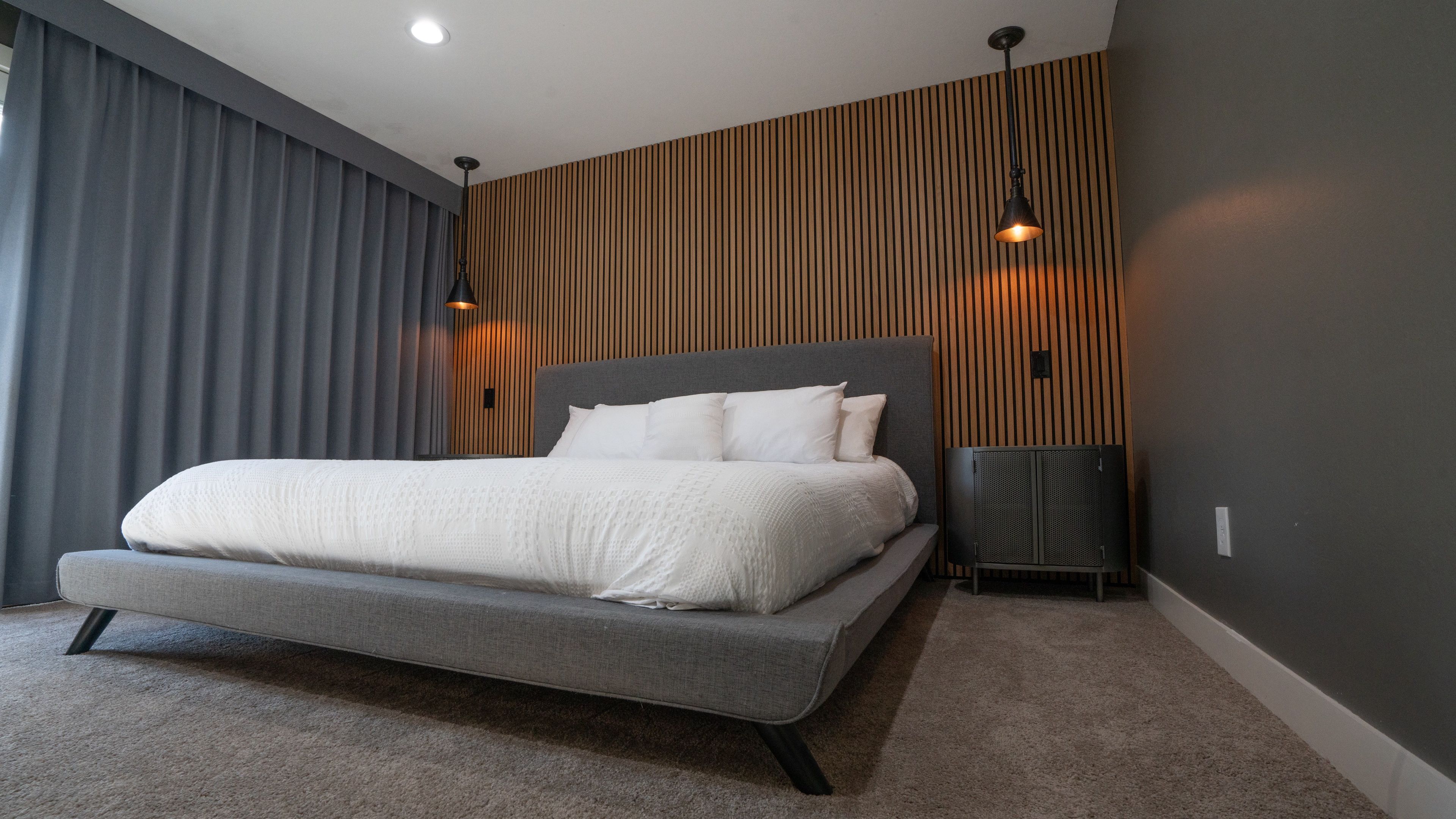 A modern bedroom with a large bed, grey walls, wooden paneling, and two pendant lights.