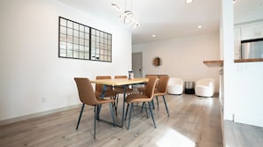 Ein moderner Essbereich mit einem Holztisch und braunen Stühlen, eine Küche mit weißen Schränken und einem großen Fenster.
