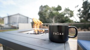 Eine schwarze Tasse mit dem Schriftzug 'relax' auf einer Metalloberfläche, im Hintergrund ein Feuerkorb.