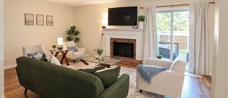 A living room with a green sofa, a white armchair, a fireplace, and a television mounted above it.
