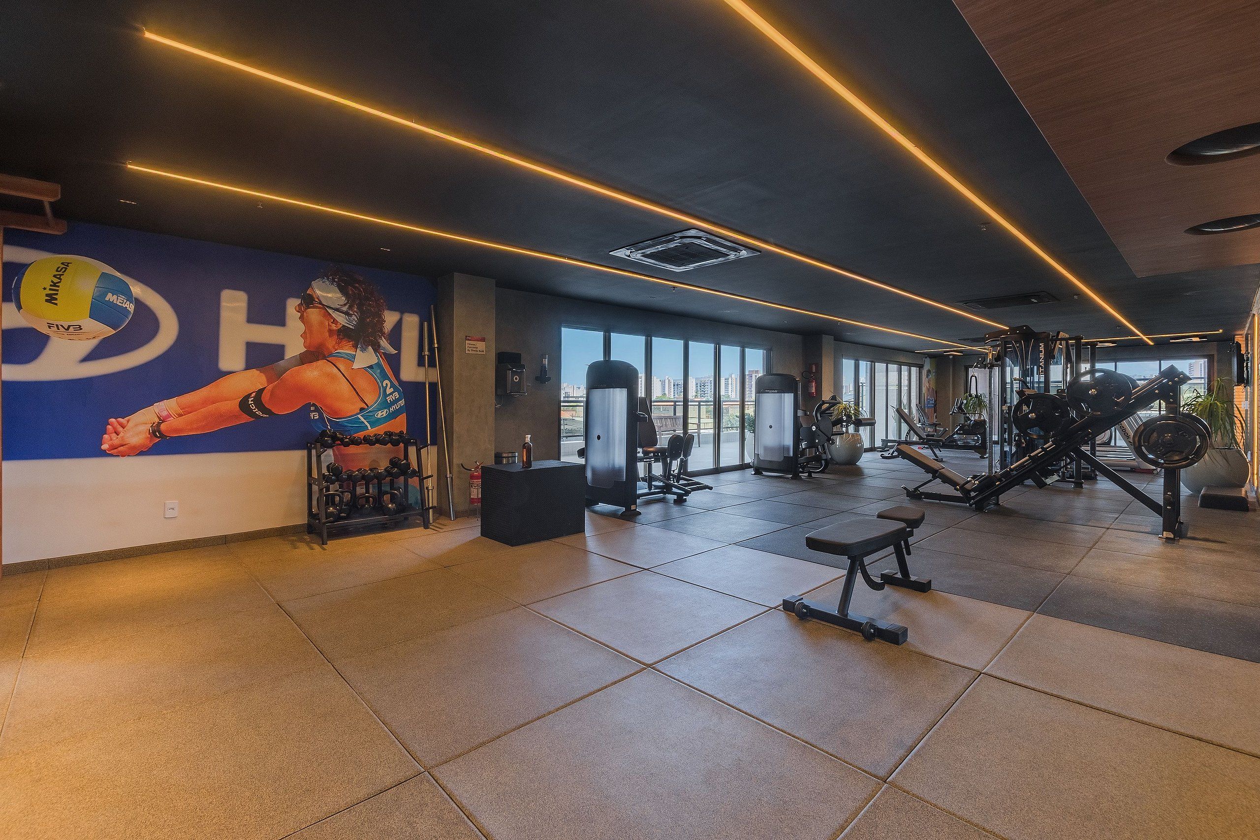 Un gimnasio con equipo de ejercicio, una pared con un jugador de voleibol y vistas a la ciudad a través de amplias ventanas.