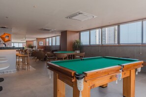 Ein Billardtisch in einem Raum mit großen Fenstern, einer Bar und Sitzgelegenheiten.