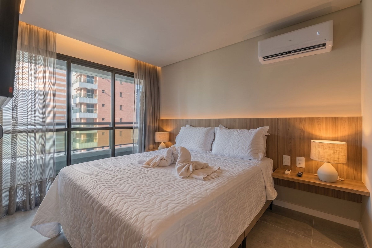 A hotel room with a bed, a nightstand, an air conditioning unit, and a view of a building through the window.