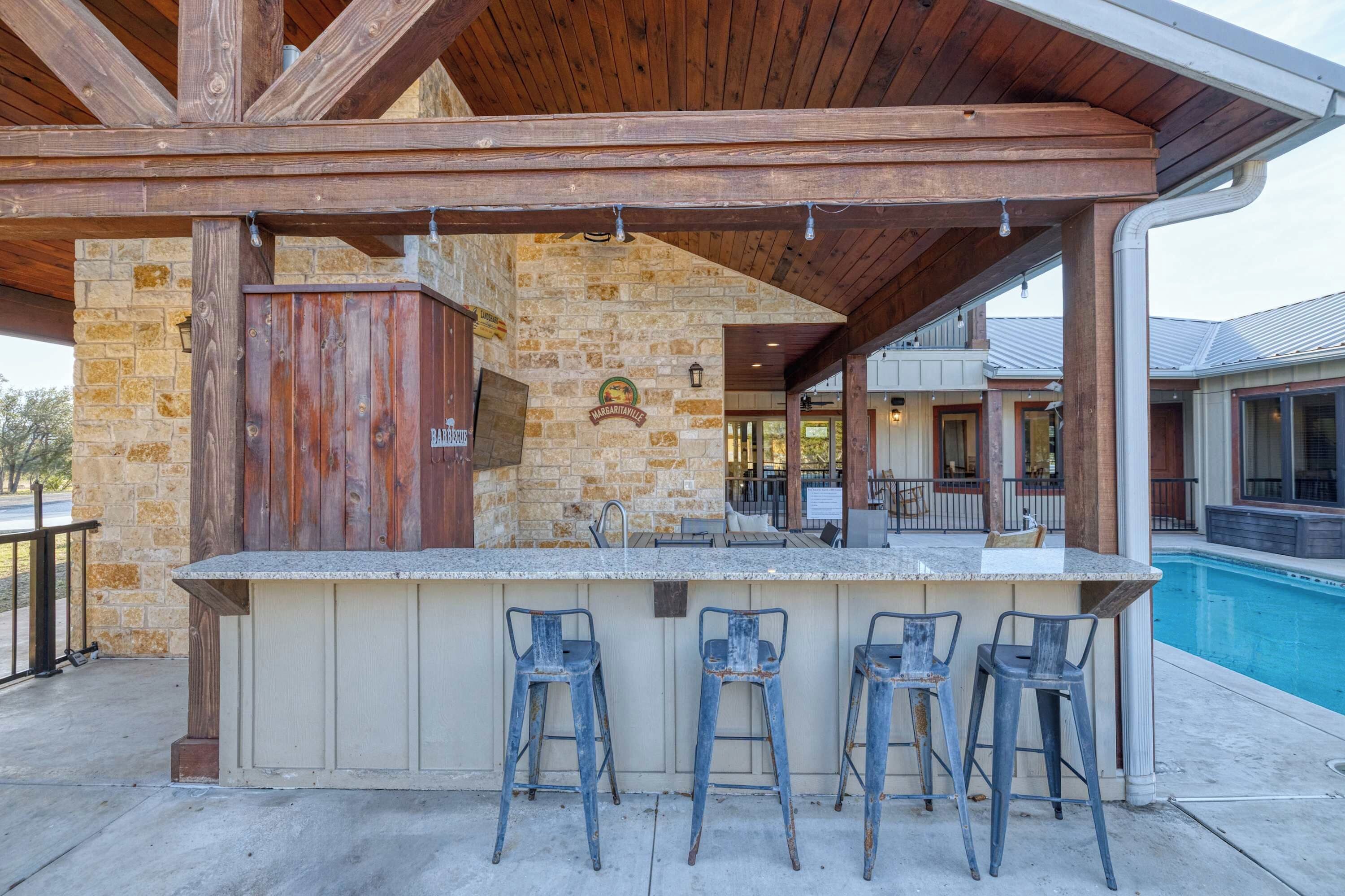 An outdoor bar area with a stone wall, wooden beams, and a pool in the background.