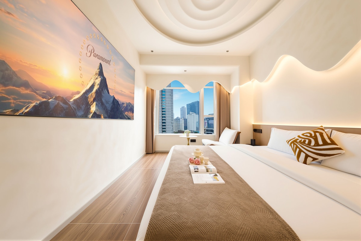 A modern hotel room with a large window, a bed, a bedside table, and a view of the cityscape.