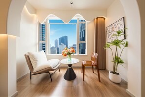 Una habitación moderna con vistas a un horizonte urbano a través de un gran ventanal, una mesa redonda con un jarrón de flores, un sofá, una silla y una planta en maceta.