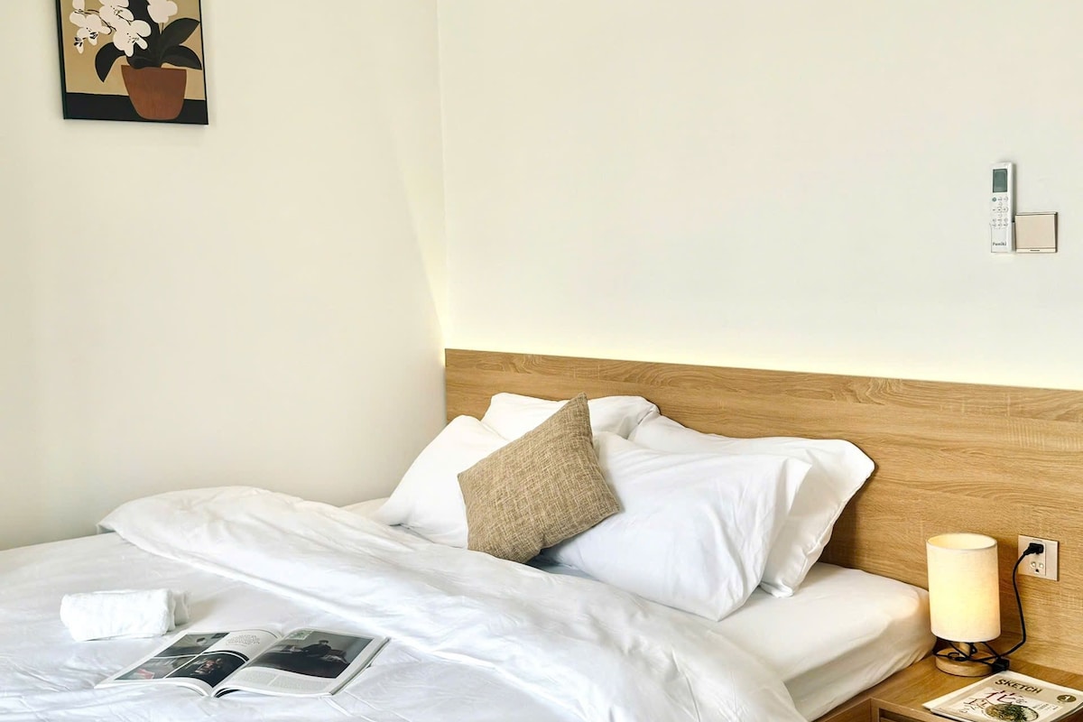 Cama bem arrumada com roupa de cama branca, cabeceira de madeira, luminária de cabeceira e uma mesinha com uma revista.