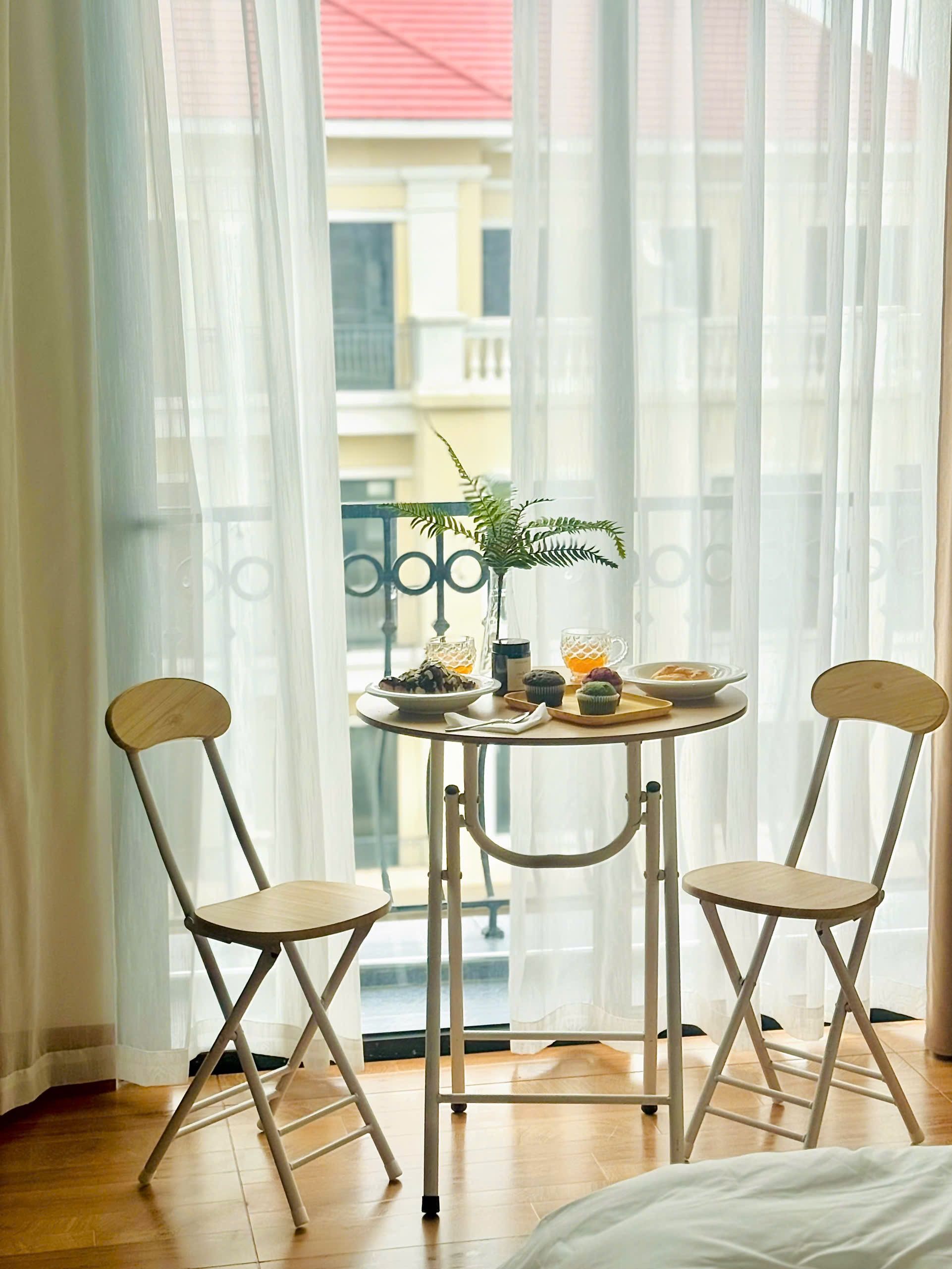 A room with a small round table, two chairs, a potted plant, and a view of buildings through sheer curtains.