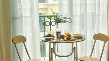 A room with a small round table, two chairs, a potted plant, and a view of buildings through sheer curtains.