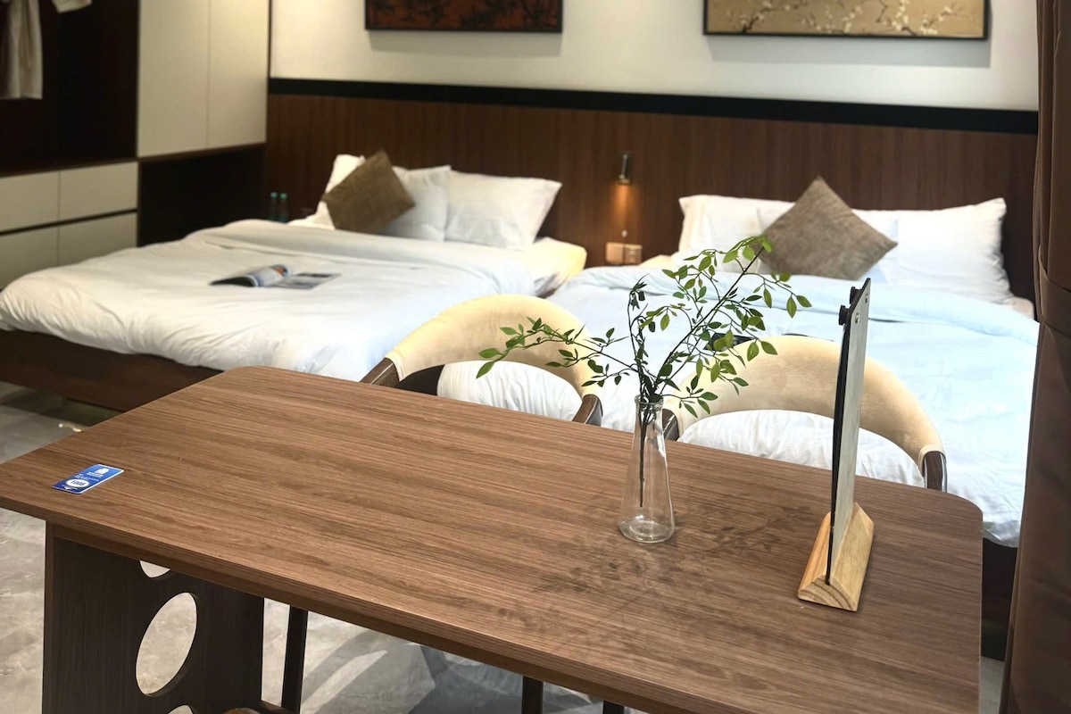 Quarto de hotel moderno com duas camas, uma mesa de madeira e um vaso com planta.